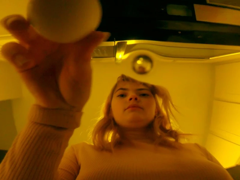 Person viewed from below holding a white egg and a metallic sphere in a warmly lit kitchen setting.