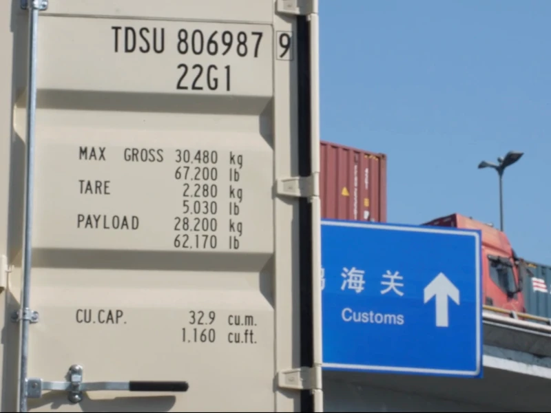Shipping container side with specifications and a nearby customs road sign.