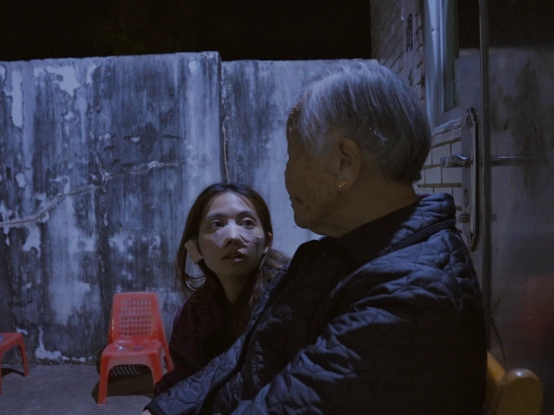 Two people sitting outside at night, one with gray hair and a quilted jacket, the other with bandages on their face. Weathered wall and red chairs in the background.