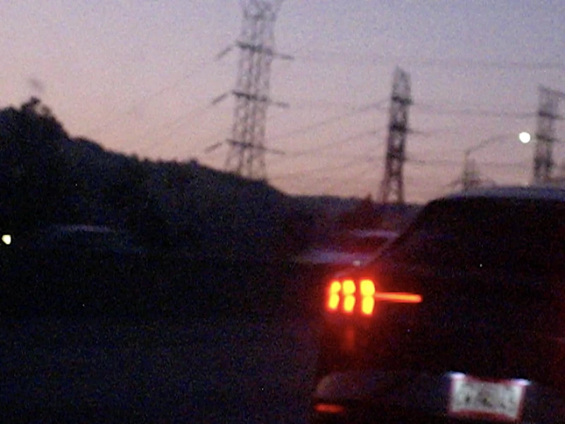 A car with bright red tail lights against a twilight sky with power lines and towers.