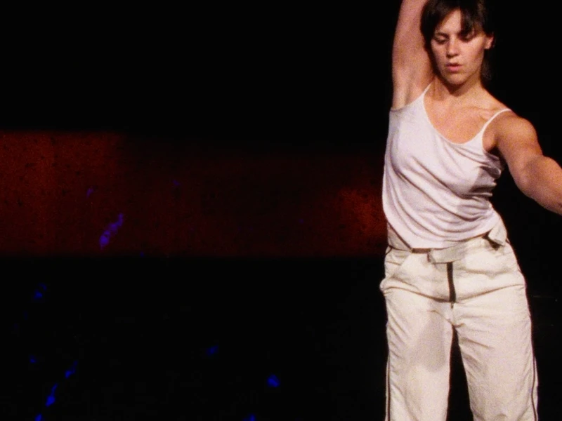 A dancer in a white tank top and light pants performs against a dark, subtly red background.