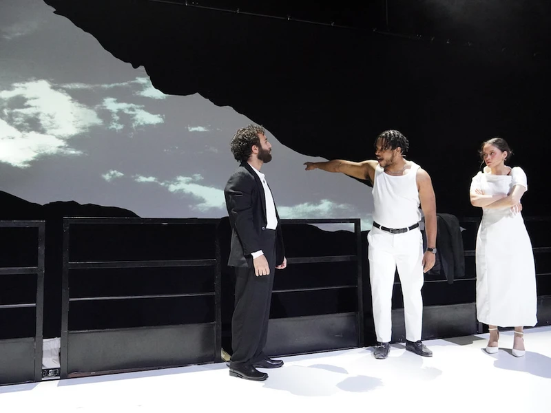 Three CalArts actors perform on stage in a production in front of a black and white projection of clouds.