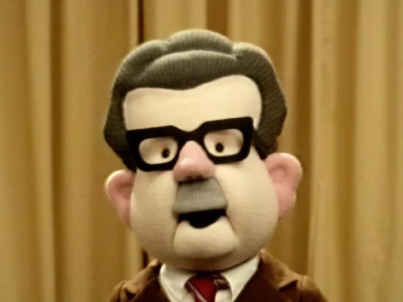 A puppet with black glasses, gray hair, and a brown suit stands in front of beige curtains.
