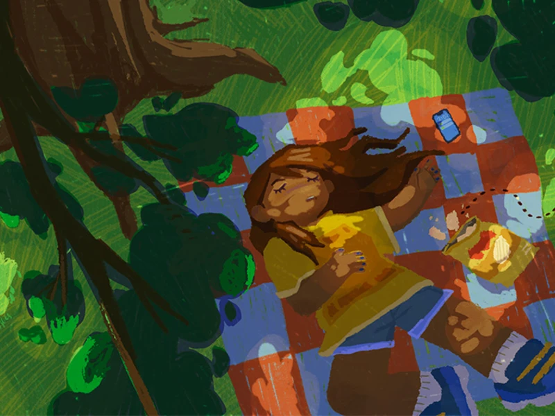Child lying on a checkered picnic blanket under trees with dappled light, surrounded by a bag of chips and a can.