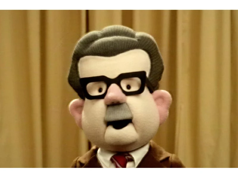 A puppet with black glasses, gray hair, and a brown suit stands in front of beige curtains.