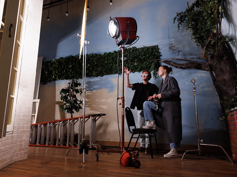 Two people in a studio setting with a large film light, surrounded by faux greenery and equipment.