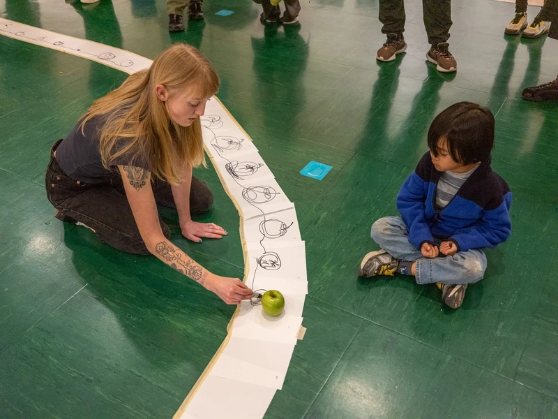 A woman drawing on paper strips with an apple sketch, a real apple nearby, and a child watching.
