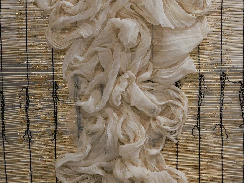 Fabric artwork with cream-colored draped fabric over bamboo sticks with gold accents and vertical black threads.