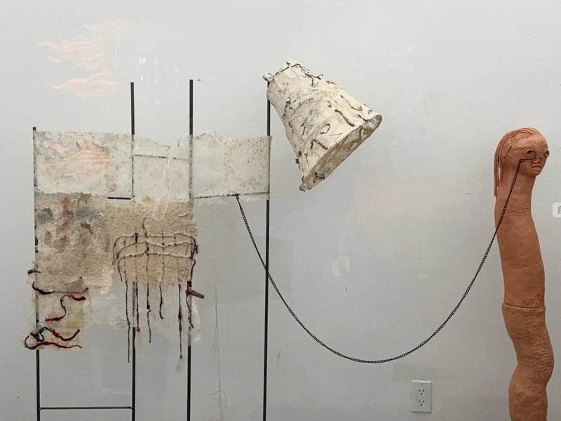 Abstract art installation with textured paper, a lampshade, and a terracotta figure linked by a chain.