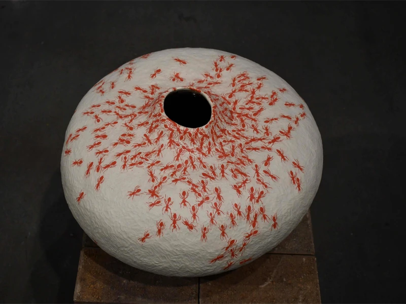 A dome-shaped sculpture with a textured off-white surface, covered in red ant figures, and a black hole at the center top.