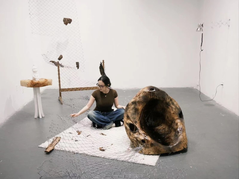 Art installation with a woman seated on a rug, examining wire mesh, a large organic sculpture, and vertical mesh with wooden fragments.