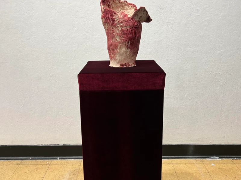 A red and ivory textured sculpture on a burgundy pedestal against a light wall.