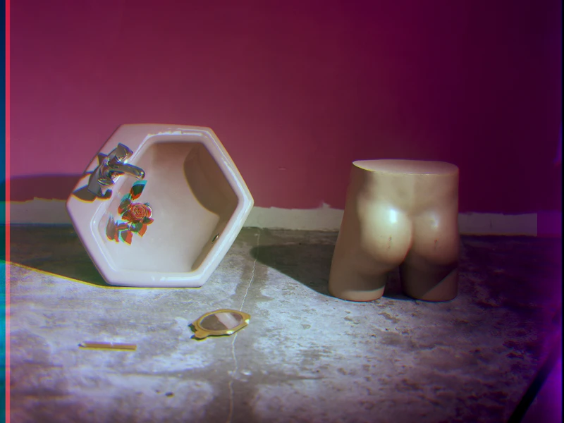 A hexagonal sink on its side beside a torso-shaped sculpture and a hand mirror on a concrete floor against a colorful wall.