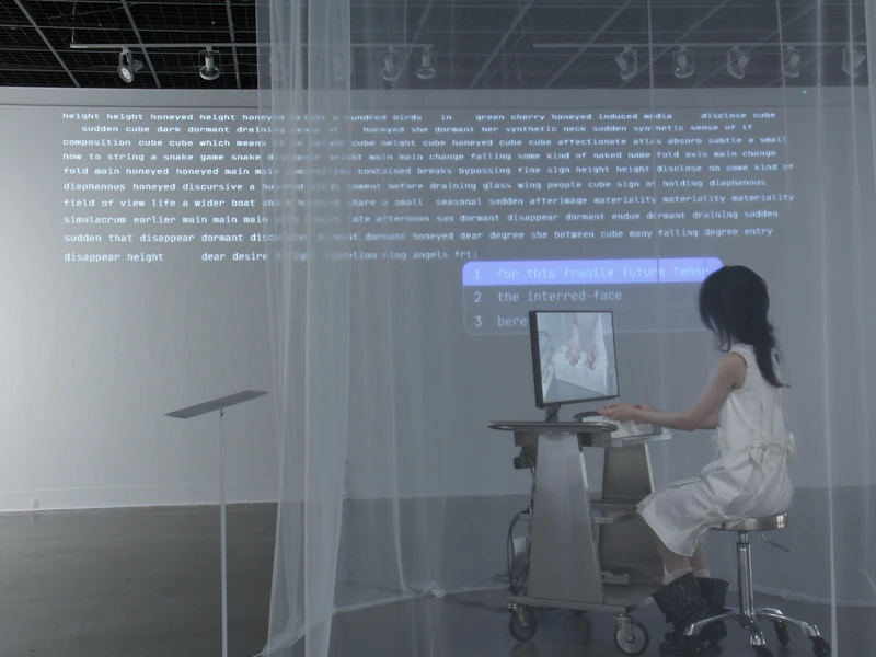 A person sitting at a desk with a computer in front of a projected wall of text in a dimly lit room with a sheer curtain.