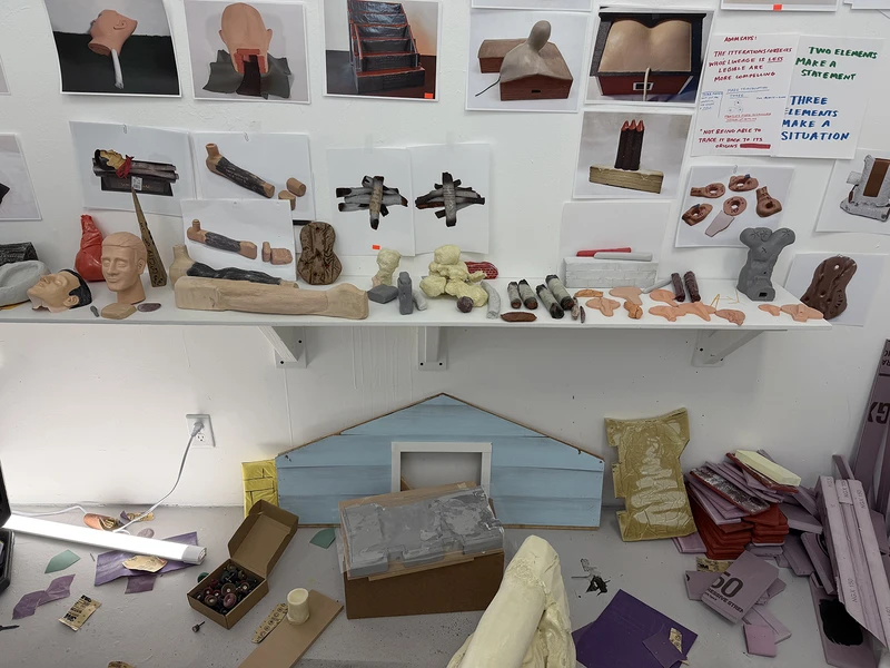 Art studio wall displaying photographs of sculptures, clay models on shelves, and scattered art supplies below.