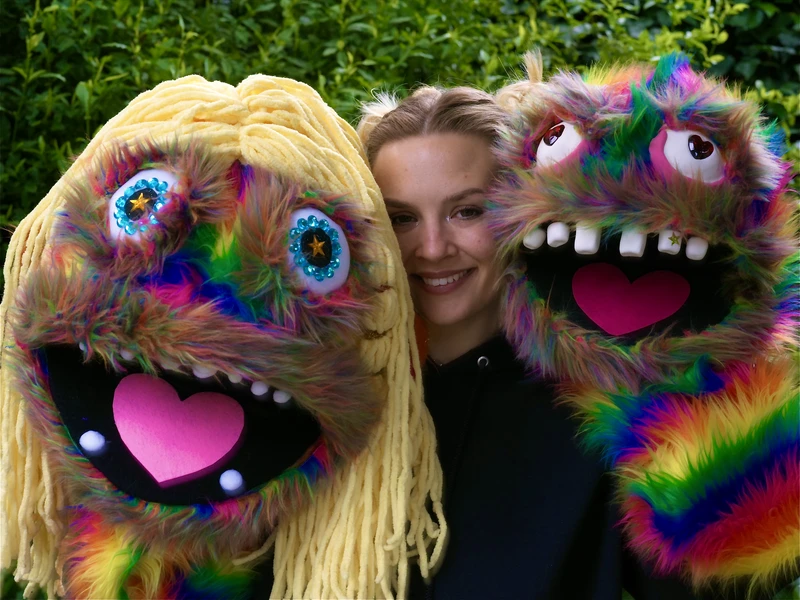 Person smiling between two large, colorful, furry puppets with rainbow patterns and playful expressions.