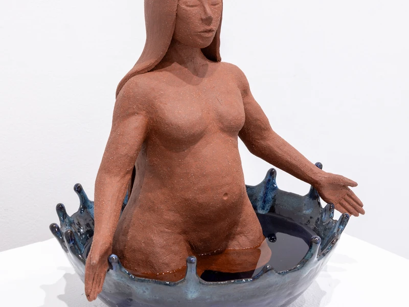 A clay sculpture of a human figure emerging from a blue, splash-shaped ceramic bowl.