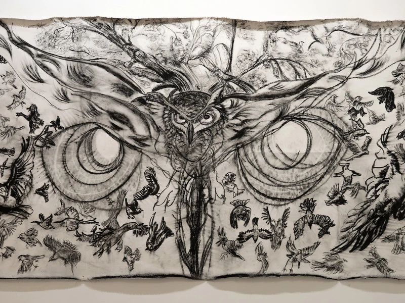 A large charcoal drawing of an owl with outstretched wings, surrounded by smaller birds in flight on a fabric canvas.