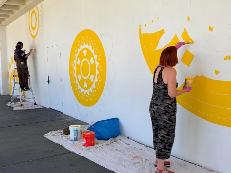 Two women are painting yellow paint on a white wall. 