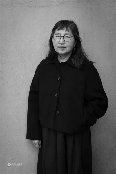 Person in a dark coat standing against a light wall.