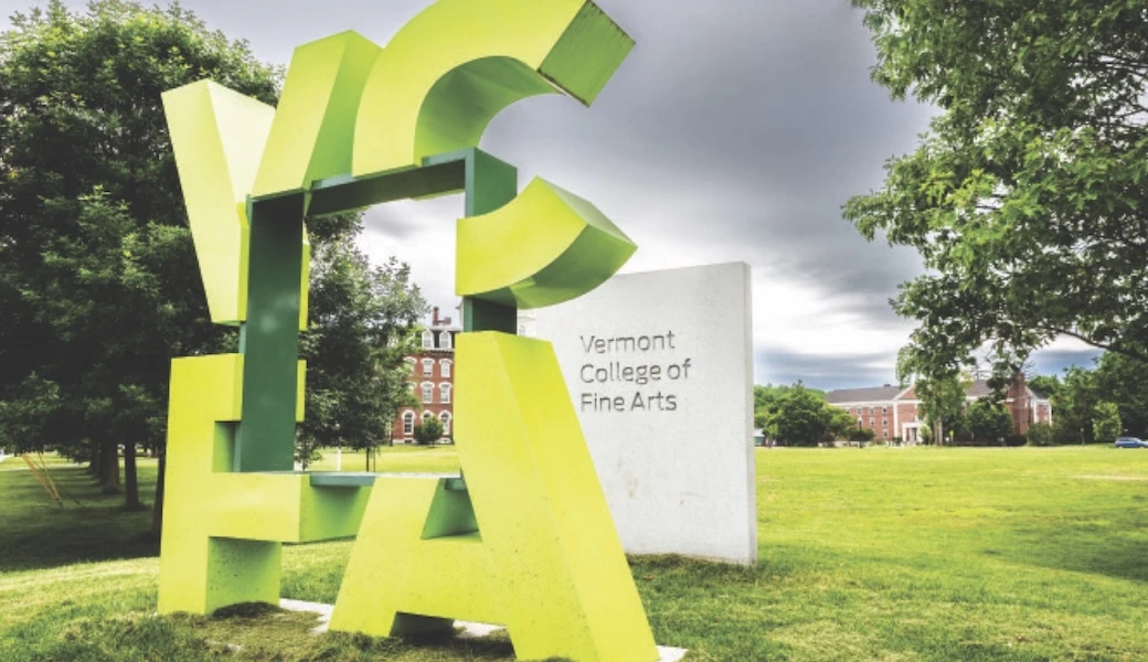 Abstract green sculpture spelling "VCFA" near Vermont College of Fine Arts sign with buildings and trees in the background.