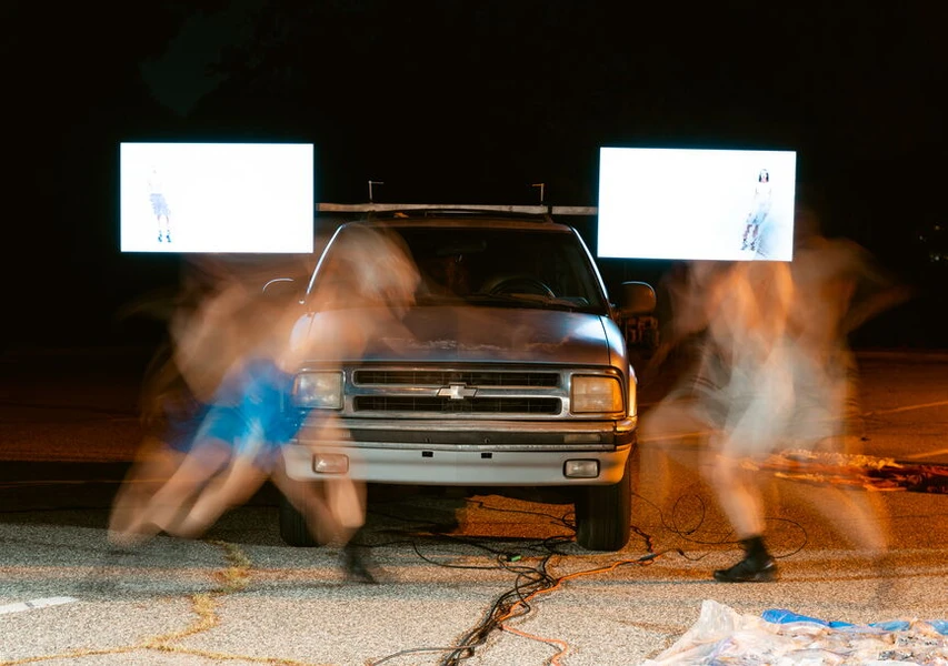 Silver Chevrolet truck outdoors at night with two bright screens and blurred figures moving in front.