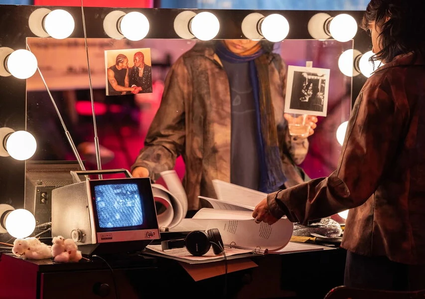 A dressing room mirror with lights, a person holding a photo, a small static-filled TV, papers, and plush toys on the table.