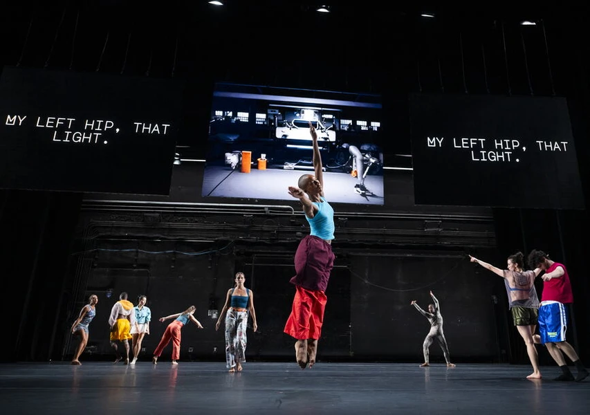 Dancers performing on stage with screens displaying the text "MY LEFT HIP, THAT LIGHT."
