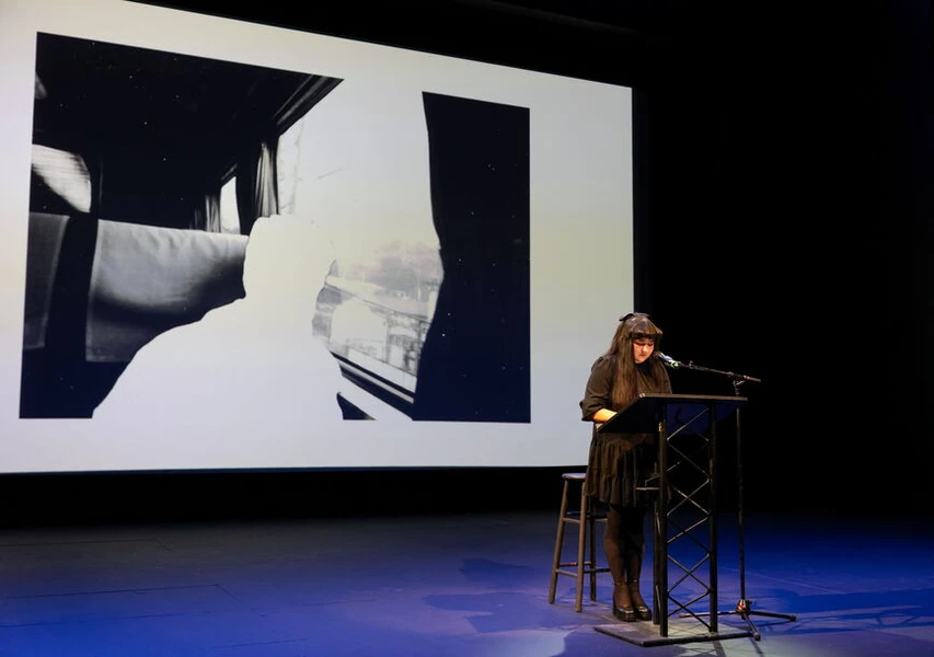 A person stands at a lectern on a stage with a black and white photo projected behind them.