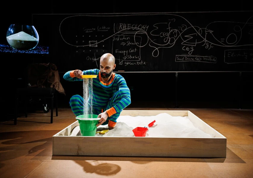 A man in a colorful striped outfit pours sand through a sieve into a green bucket in a sandbox, with a blackboard covered in scientific drawings behind him.