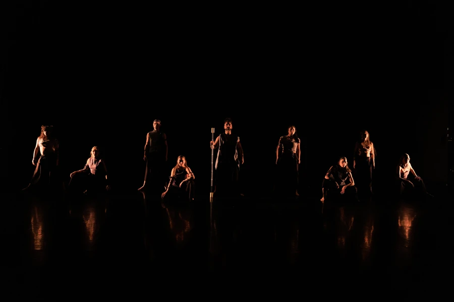 A group of performers on a dimly lit stage, with dramatic lighting casting reflections on the floor.