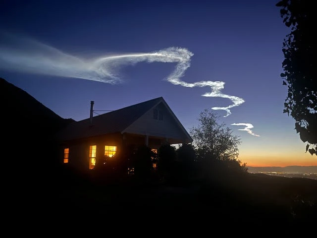 A house silhouetted against a sunset sky with winding white clouds and illuminated windows.