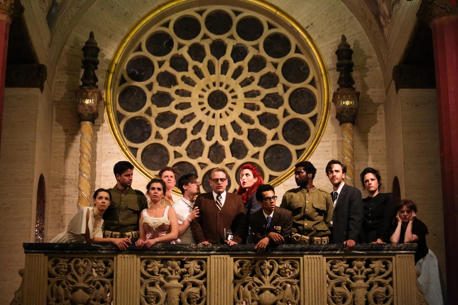 A group of people stands on a decorative balcony inside a historic building, with a large stained-glass window behind them.
