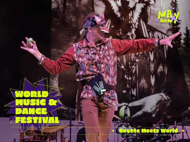 A performer at the World Music & Dance Festival wearing a coyote mask holds a green ball on stage with a coyote image in the background.