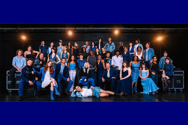 A diverse group of people in denim clothing posed on a stage with overhead lights.