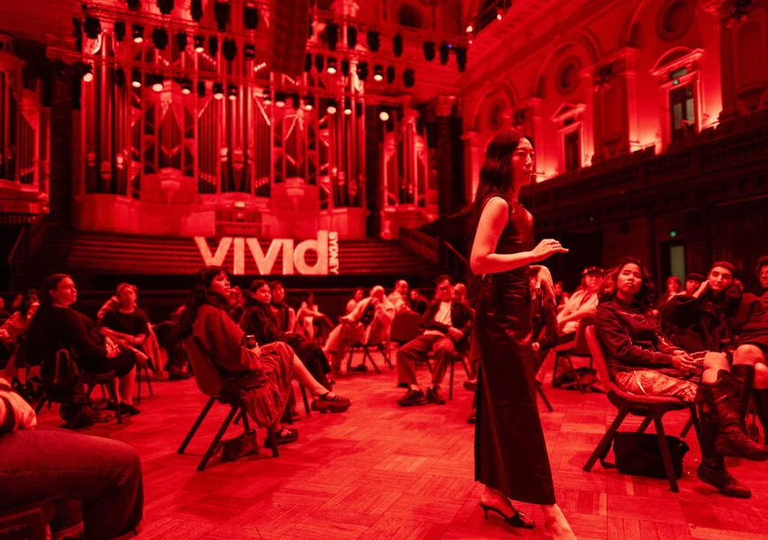 An engaging speaker in a black dress stands in front of an audience in a red-lit hall, with diverse attendees seated in a semicircle and the word "vivid" displayed in white behind them.