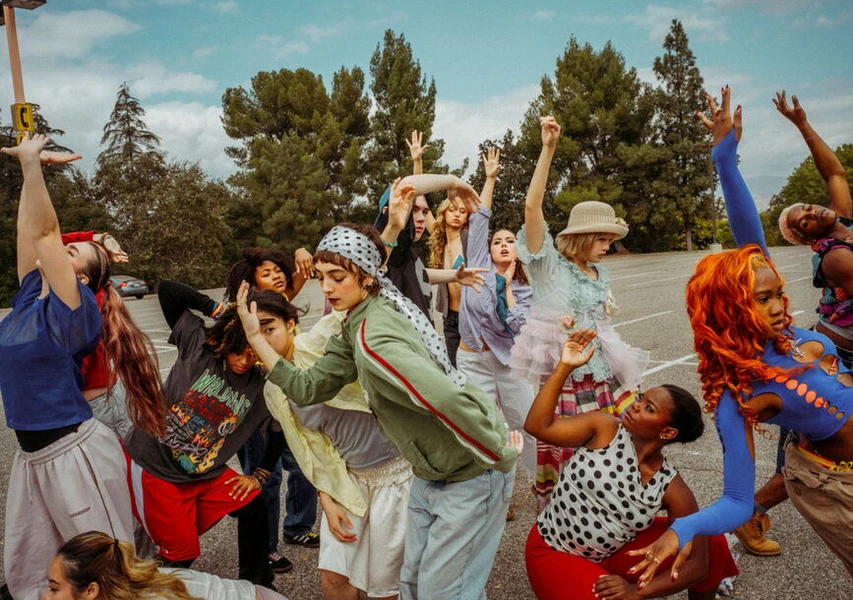 A diverse group of individuals poses energetically in an outdoor parking lot, showcasing various fashion styles against a backdrop of trees and clouds.