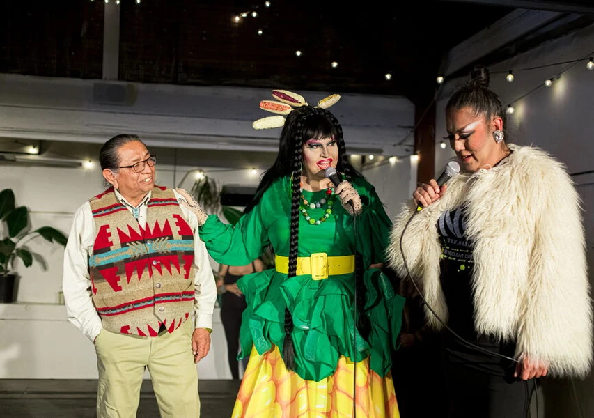 A drag performer in a green leaf-inspired dress and a man in a colorful vest are on stage, interacting with another performer in a white coat. String lights illuminate the backdrop.