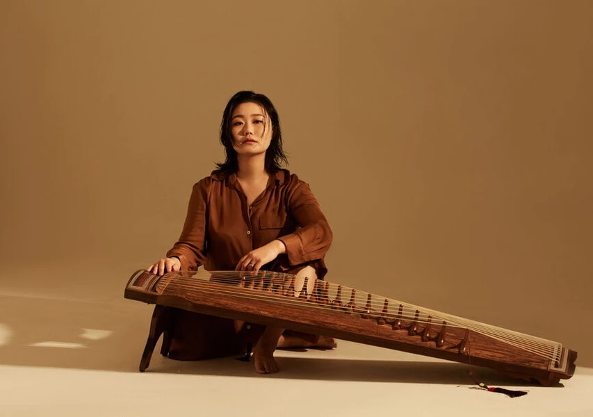 A woman seated cross-legged on a light floor, playing a traditional zither-like instrument, dressed in a brown shirt against a beige background.