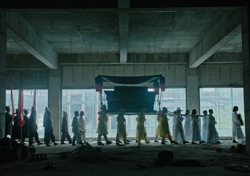 A procession of individuals in various clothing, carrying a large, draped object through a dimly lit industrial space with concrete walls.