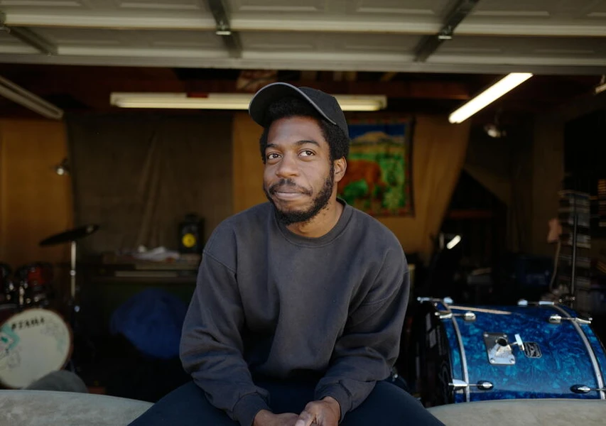 A young Black man in a black cap and sweatshirt sits in a sunlit garage among musical instruments, smiling slightly.