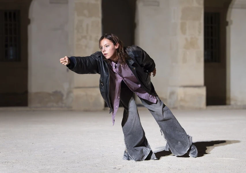 A young woman in a black leather jacket and frayed gray pants stands in an expressive pose, extending her arm forward, against a softly lit stone background.