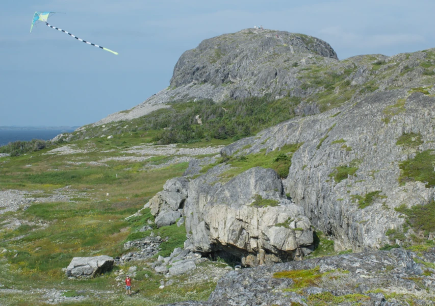 A colorful kite flies above a rocky hill as a person in a red shirt stands nearby on a grassy landscape.