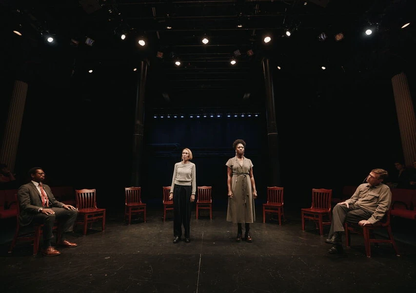 Two female actors stand on stage, flanked by red wooden chairs, with two male actors seated. The scene is lit dramatically against a dark background.
