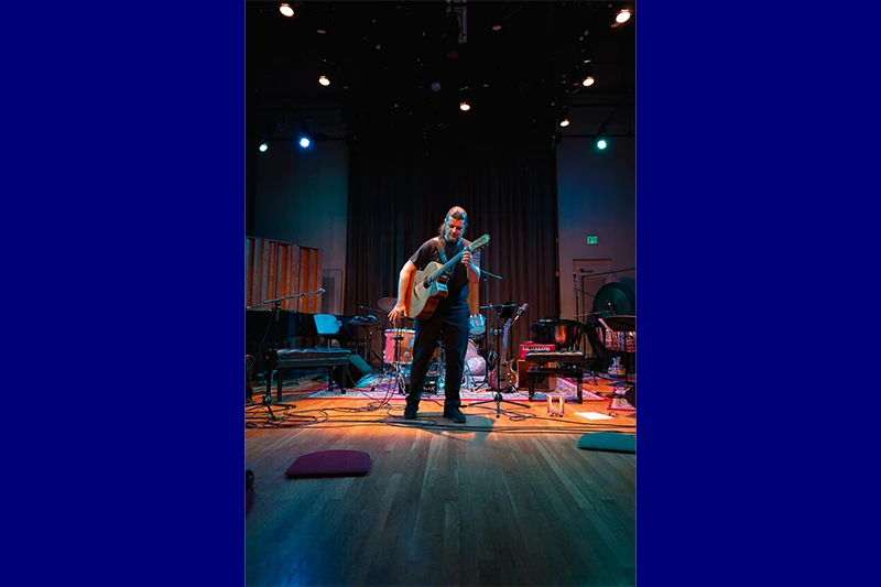 A musician stands on stage with an acoustic guitar, surrounded by various musical instruments, under colorful stage lights.