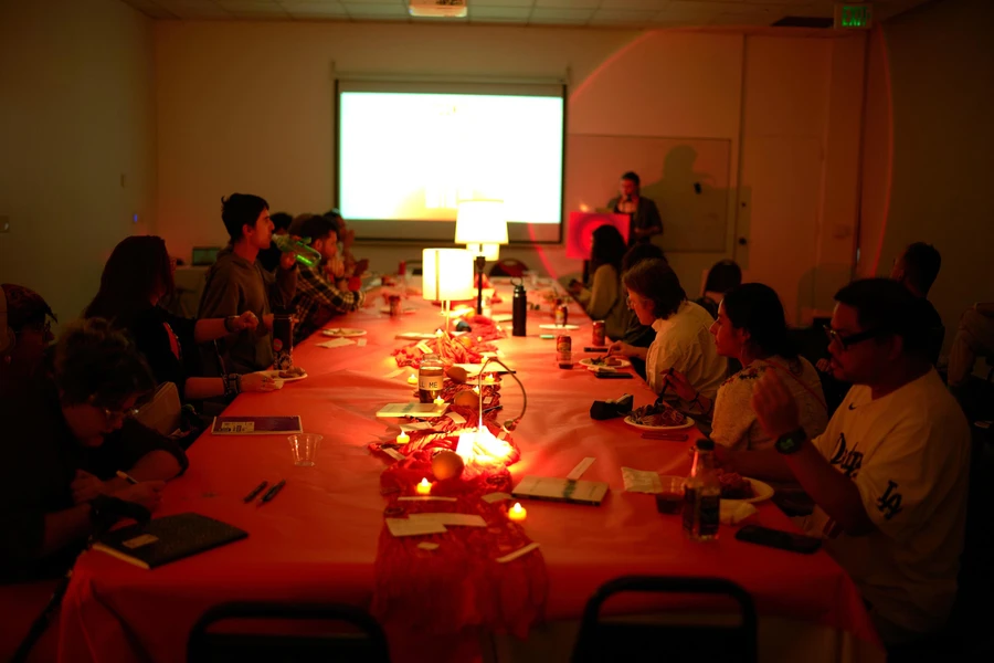 People dine around a long red-covered table while a speaker stands beside a projection screen in a dim, red-lit room.