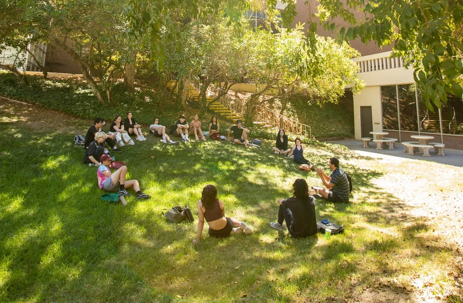 Group of people sitting on a shaded grassy courtyard beside a brick-and-cream building with stairs and picnic tables.