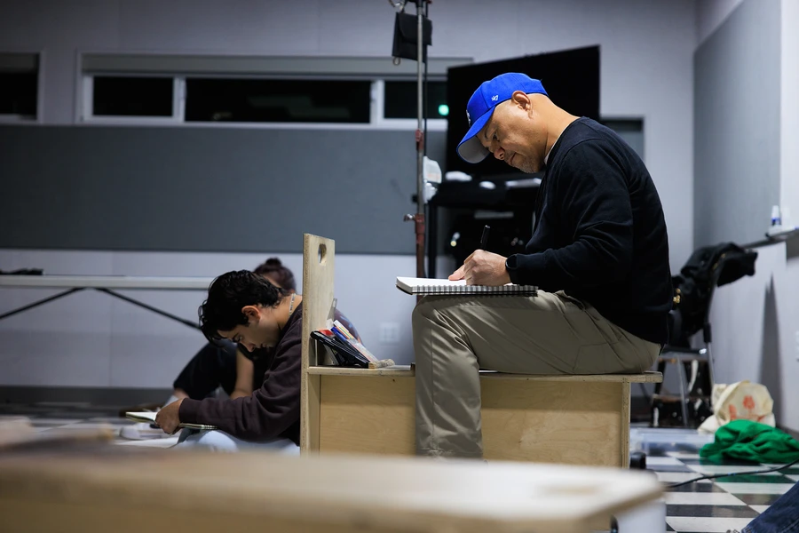 Two people work in notebooks in a studio, with one seated on a wooden platform and writing in a sketchbook.
